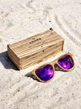 Load image into Gallery viewer, Zebrawood wooden sunglasses with purple polarized lenses and a box on sandy beach.