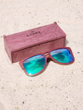 Load image into Gallery viewer, Purpleheart wooden sunglasses with green polarized lenses and a box on sandy beach.