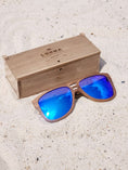 Load image into Gallery viewer, Walnut wooden sunglasses with blue polarized lenses and a box on sandy beach.