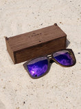 Load image into Gallery viewer, Wenge wooden sunglasses with purple polarized lenses and a box on sandy beach.