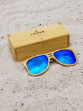 Load image into Gallery viewer, Yellowheart wooden sunglasses with blue polarized lenses and a box on sandy beach.