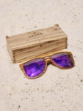 Load image into Gallery viewer, Zebrawood wooden sunglasses with purple polarized lenses and a box on sandy beach.