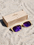 Load image into Gallery viewer, Maple wooden sunglasses with purple polarized lenses and a box on sandy beach.