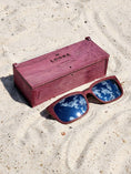 Load image into Gallery viewer, Purpleheart wooden sunglasses with silver polarized lenses and a box on sandy beach.