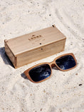 Load image into Gallery viewer, Walnut wooden sunglasses with black polarized lenses and a box on sandy beach.
