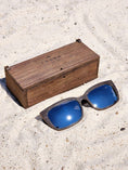 Load image into Gallery viewer, Wenge wooden sunglasses with silver polarized lenses and a box on sandy beach.