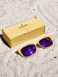 Load image into Gallery viewer, Yellowheart wooden sunglasses with purple polarized lenses and a box on sandy beach.