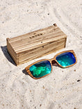 Load image into Gallery viewer, Zebrawood wooden sunglasses with green polarized lenses and a box on sandy beach.