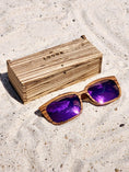 Load image into Gallery viewer, Zebrawood wooden sunglasses with purple polarized lenses and a box on sandy beach.