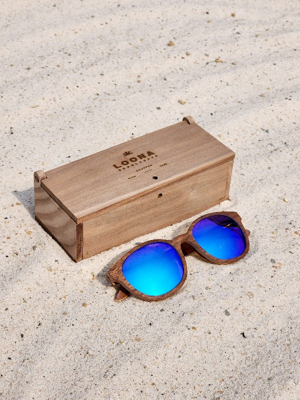 Walnut wooden sunglasses with blue polarized lenses and a box on sandy beach.