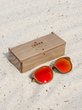 Load image into Gallery viewer, Walnut wooden sunglasses with red polarized lenses and a box on sandy beach.

