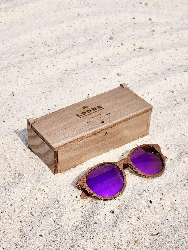 Walnut wooden sunglasses with purple polarized lenses and a box on sandy beach.