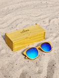 Load image into Gallery viewer, Yellowheart wooden sunglasses with blue polarized lenses and a box on sandy beach.
