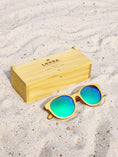 Load image into Gallery viewer, Yellowheart wooden sunglasses with green polarized lenses and a box on sandy beach.
