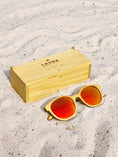 Load image into Gallery viewer, Yellowheart wooden sunglasses with red polarized lenses and a box on sandy beach.
