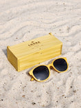 Load image into Gallery viewer, Yellowheart wooden sunglasses with black polarized lenses and a box on sandy beach.
