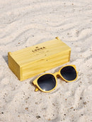 Yellowheart wooden sunglasses with black polarized lenses and a box on sandy beach.