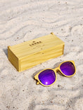 Load image into Gallery viewer, Yellowheart wooden sunglasses with purple polarized lenses and a box on sandy beach.
