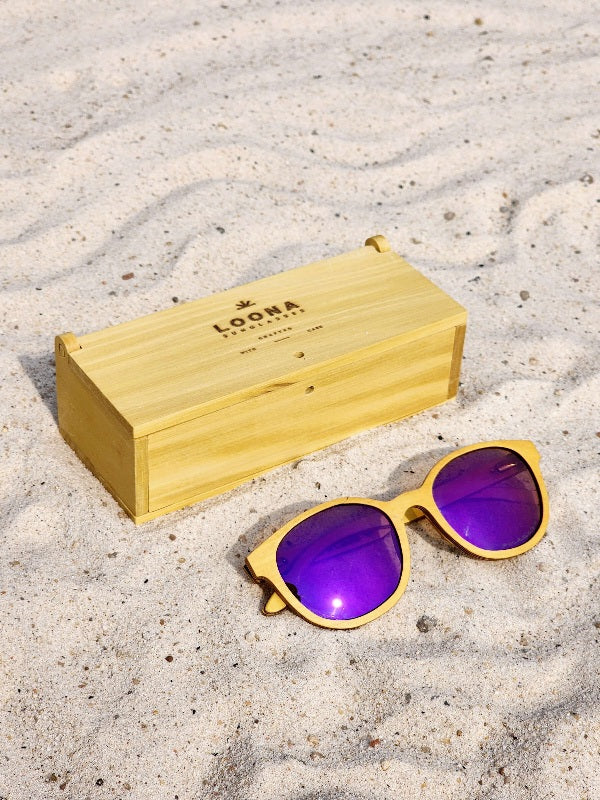Yellowheart wooden sunglasses with purple polarized lenses and a box on sandy beach.