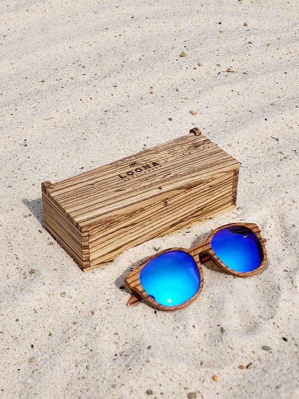 Zebrawood wooden sunglasses with blue polarized lenses and a box on sandy beach.