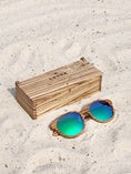 Load image into Gallery viewer, Zebrawood wooden sunglasses with green polarized lenses and a box on sandy beach.
