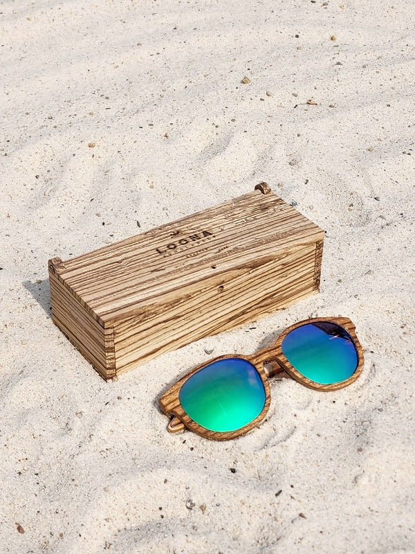 Zebrawood wooden sunglasses with green polarized lenses and a box on sandy beach.