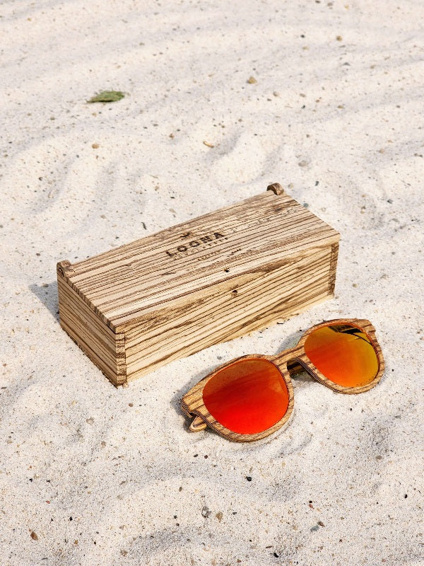 Zebrawood wooden sunglasses with red polarized lenses and a box on sandy beach.