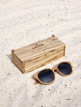 Load image into Gallery viewer, Zebrawood wooden sunglasses with black polarized lenses and a box on sandy beach.
