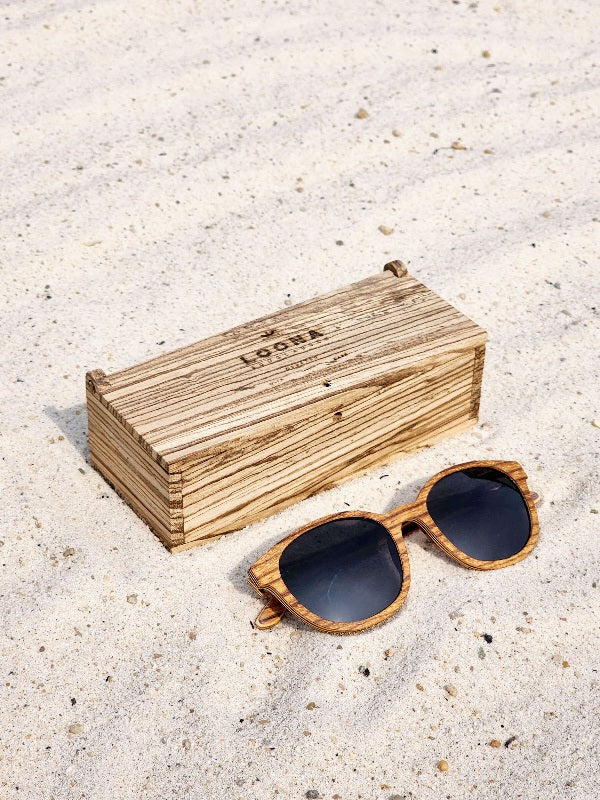 Zebrawood wooden sunglasses with black polarized lenses and a box on sandy beach.