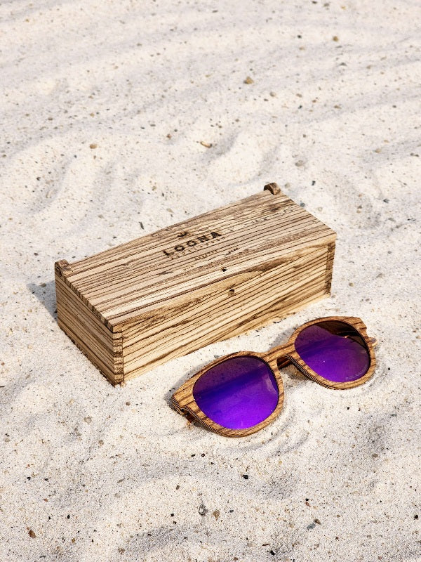 Zebrawood wooden sunglasses with purple polarized lenses and a box on sandy beach.