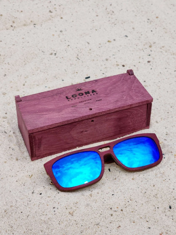 Purpleheart wooden sunglasses with blue polarized lenses and a box on sandy beach.