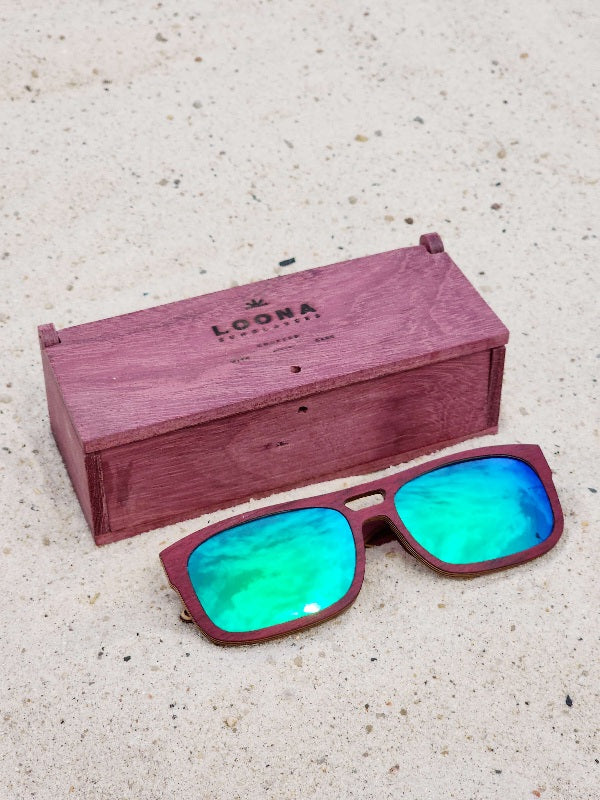 Purpleheart wooden sunglasses with green polarized lenses and a box on sandy beach.
