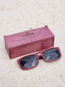 Purpleheart wooden sunglasses with black polarized lenses and a box on sandy beach.