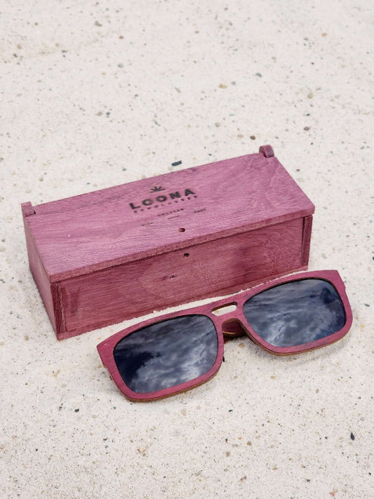 Purpleheart wooden sunglasses with black polarized lenses and a box on sandy beach.