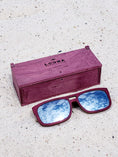 Load image into Gallery viewer, Purpleheart wooden sunglasses with silver polarized lenses and a box on sandy beach.
