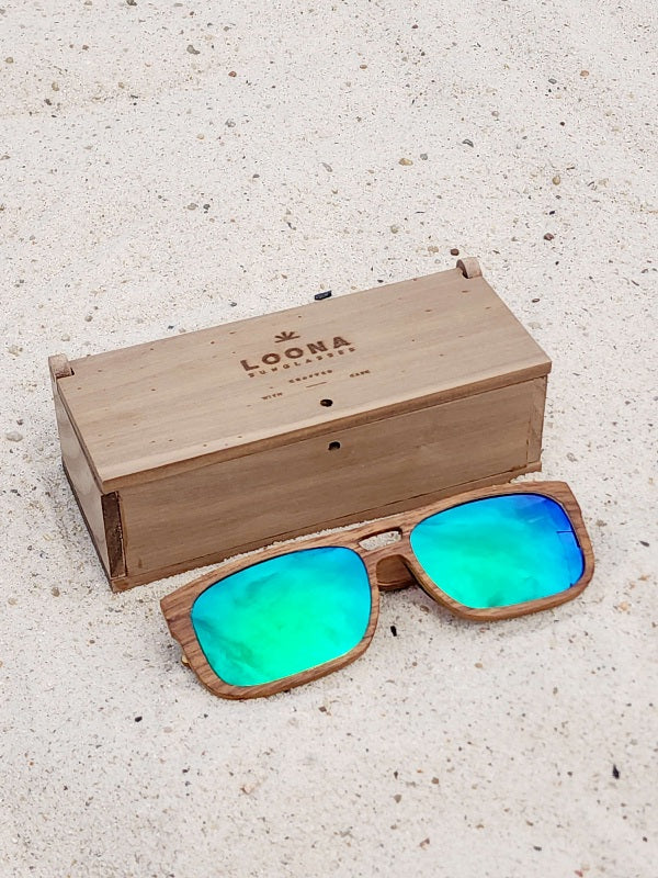 Walnut wooden sunglasses with green polarized lenses and a box on sandy beach.