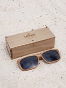 Walnut wooden sunglasses with black polarized lenses and a box on sandy beach.
