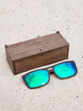 Load image into Gallery viewer, Wenge wooden sunglasses with green polarized lenses and a box on sandy beach.
