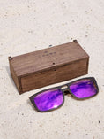 Load image into Gallery viewer, Wenge wooden sunglasses with purple polarized lenses and a box on sandy beach.
