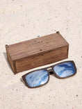 Load image into Gallery viewer, Wenge wooden sunglasses with silver polarized lenses and a box on sandy beach.

