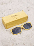 Load image into Gallery viewer, Yellowheart wooden sunglasses with black polarized lenses and a box on sandy beach.
