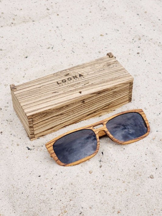 Zebrawood wooden sunglasses with black polarized lenses and a box on sandy beach.