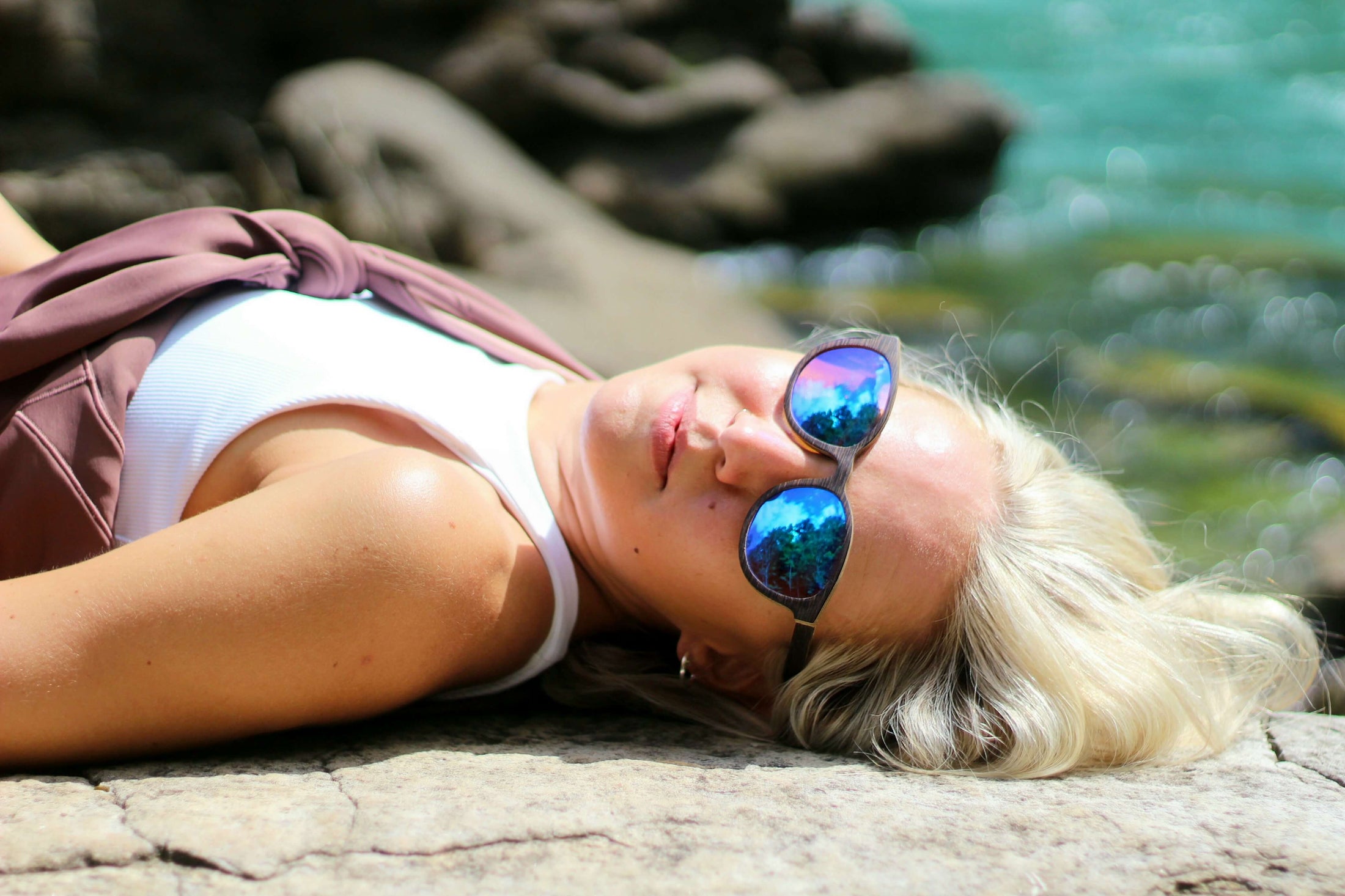 A woman laying on a rock, wearing wooden sunglasses, enjoying the serene surroundings.