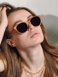 Load image into Gallery viewer,  A stylish woman wearing Zebrawood wooden sunglasses with a gold chain around her neck.
