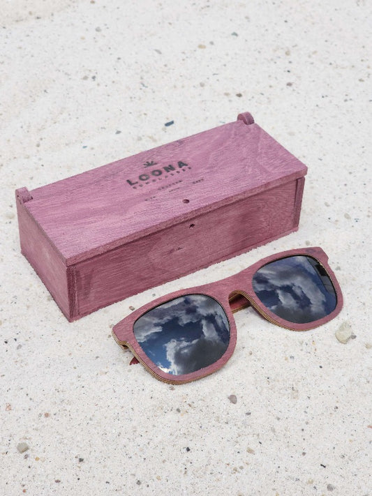 Purpleheart wooden sunglasses with black polarized lenses and a box on sandy beach.