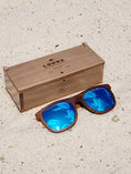 Load image into Gallery viewer, Walnut wooden sunglasses with blue polarized lenses and a box on sandy beach.
