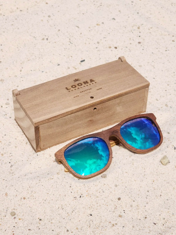 Walnut wooden sunglasses with green polarized lenses and a box on sandy beach.
