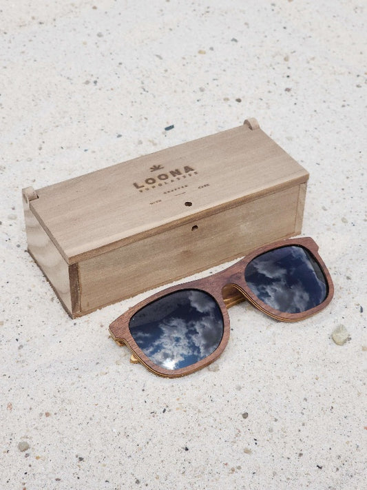 Walnut wooden sunglasses with black polarized lenses and a box on sandy beach.