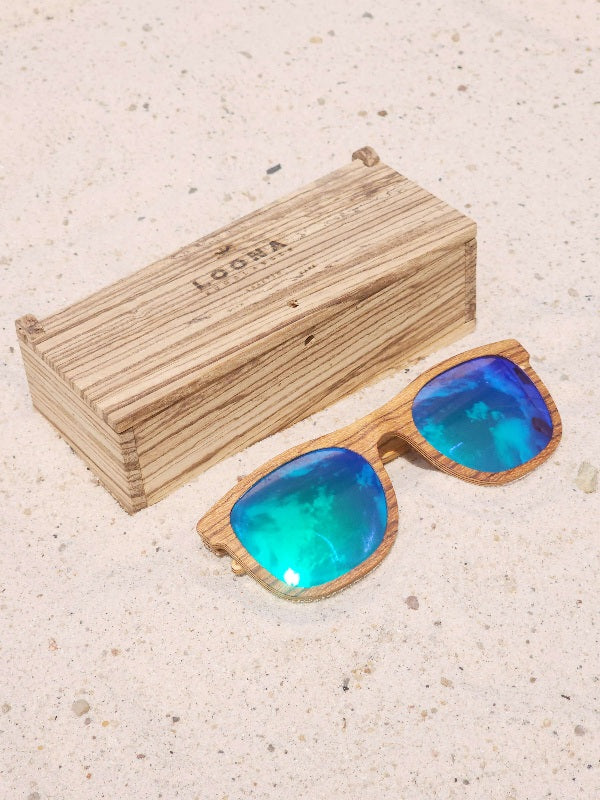 Zebrawood wooden sunglasses with green polarized lenses and a box on sandy beach.