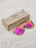 Load image into Gallery viewer, Zebrawood wooden sunglasses with red polarized lenses and a box on sandy beach.
